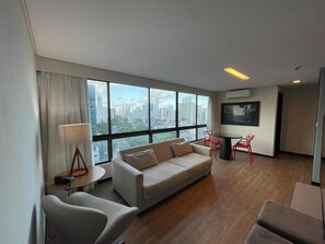 A modern hotel room with a large window offering a city view, a sofa, a small table with a lamp, and a red chair. - Flat Aconchegante em Boa Viagem (Recife)