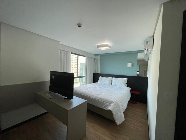 A modern hotel room with a bed, a TV on a stand, a small table, and a window with curtains. - Flat Aconchegante em Boa Viagem (Recife)