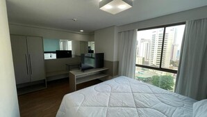 A modern hotel room with a large bed, a desk with a laptop, a television, and a window with a city view. - Flat Aconchegante em Boa Viagem (Recife)