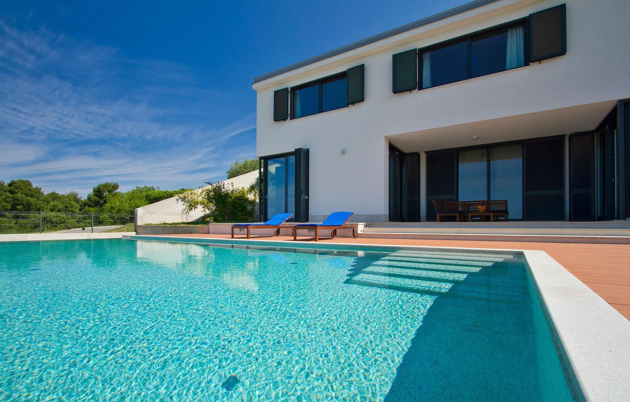 A modern two-story house with a swimming pool, blue lounge chairs, and a patio area.