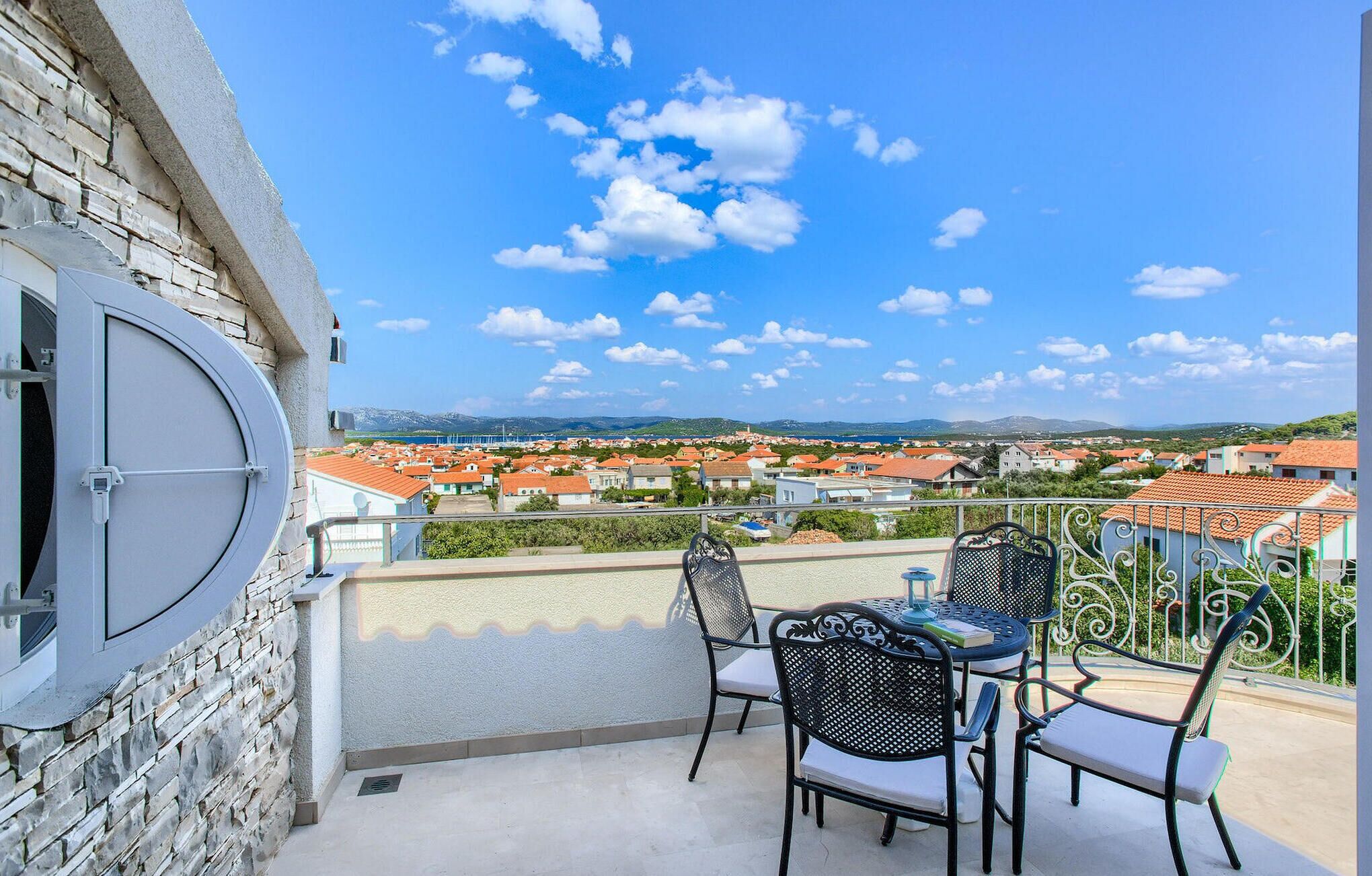 A balcony with a view of a coastal town, featuring a stone wall, metal railing, and outdoor furniture.