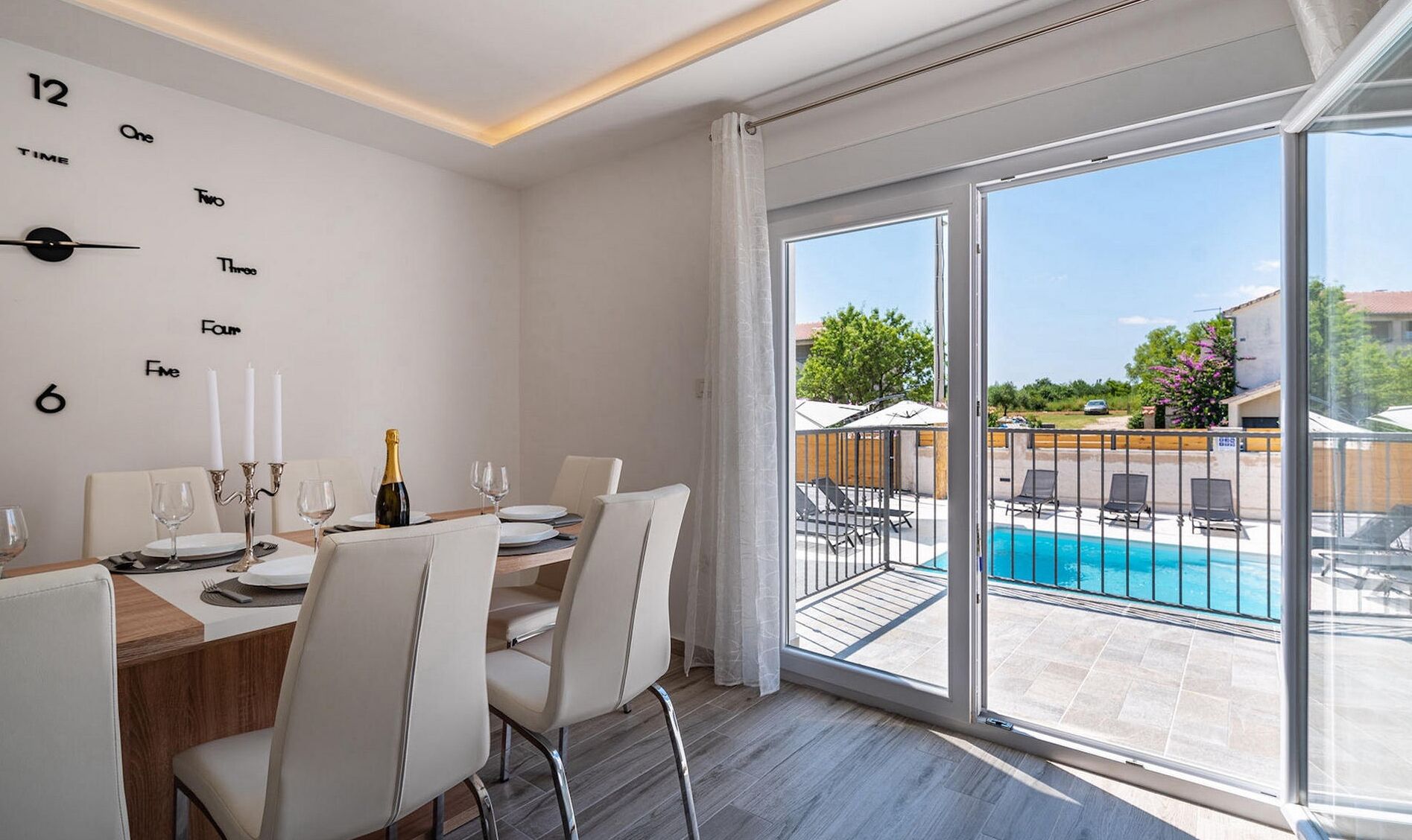 Un comedor con una mesa preparada para cuatro personas, una botella de champán y vistas a una piscina y zona de estar exterior.