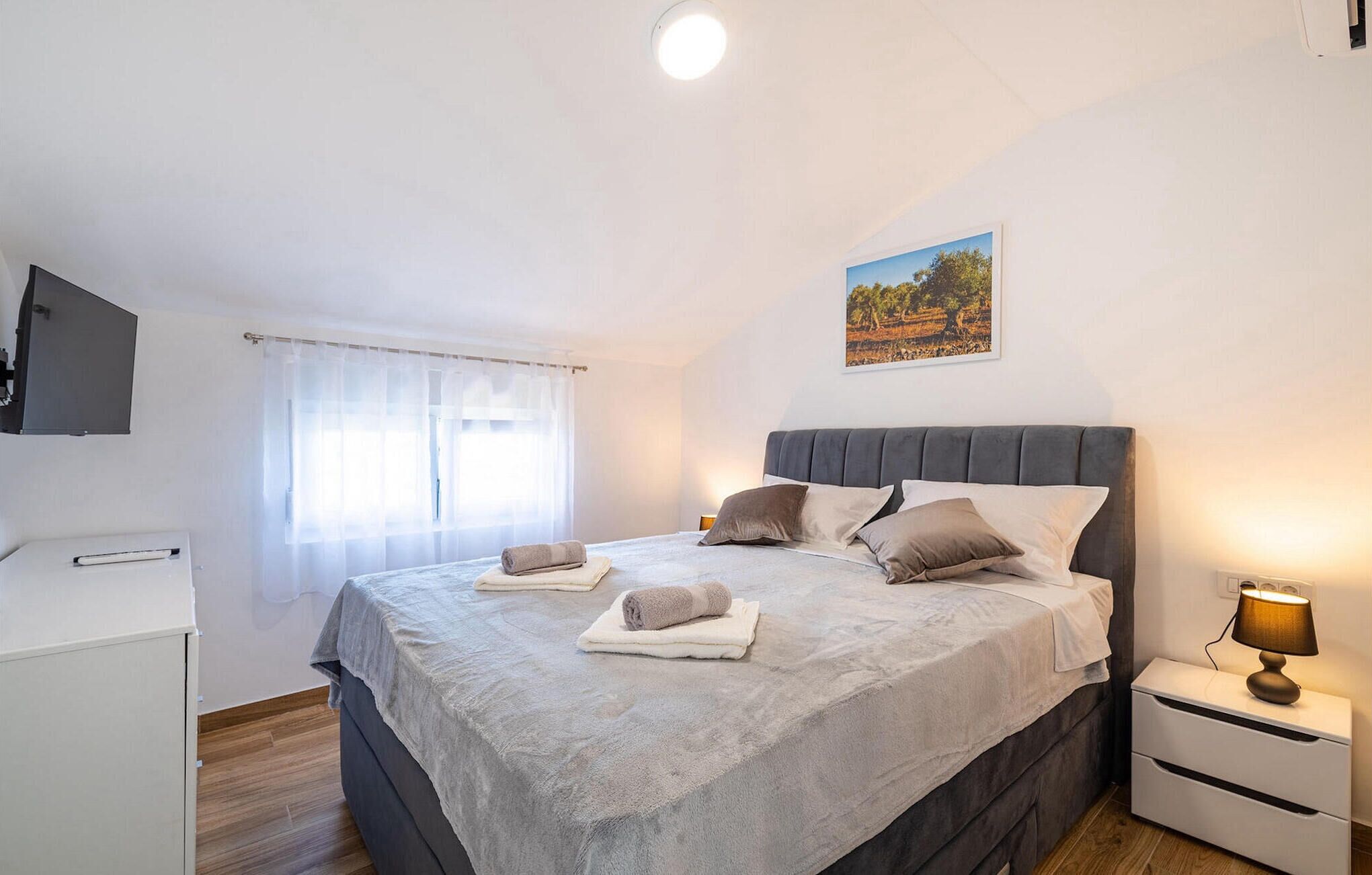 Une chambre à coucher avec un lit, une table de chevet avec une lampe, un téléviseur fixé au mur et une fenêtre avec des rideaux légers.