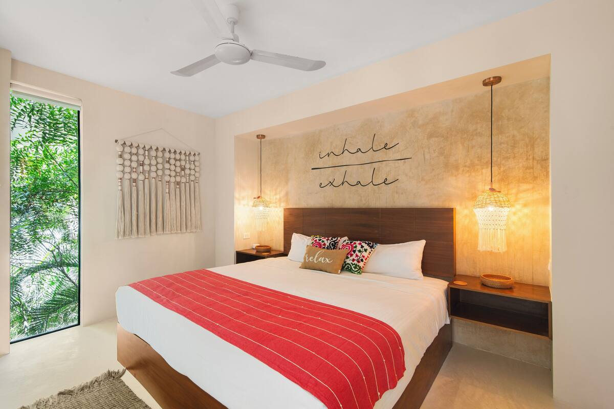 A bedroom with a bed, a ceiling fan, wall-mounted lights, a large window with a view of trees, and a motivational quote on the wall.