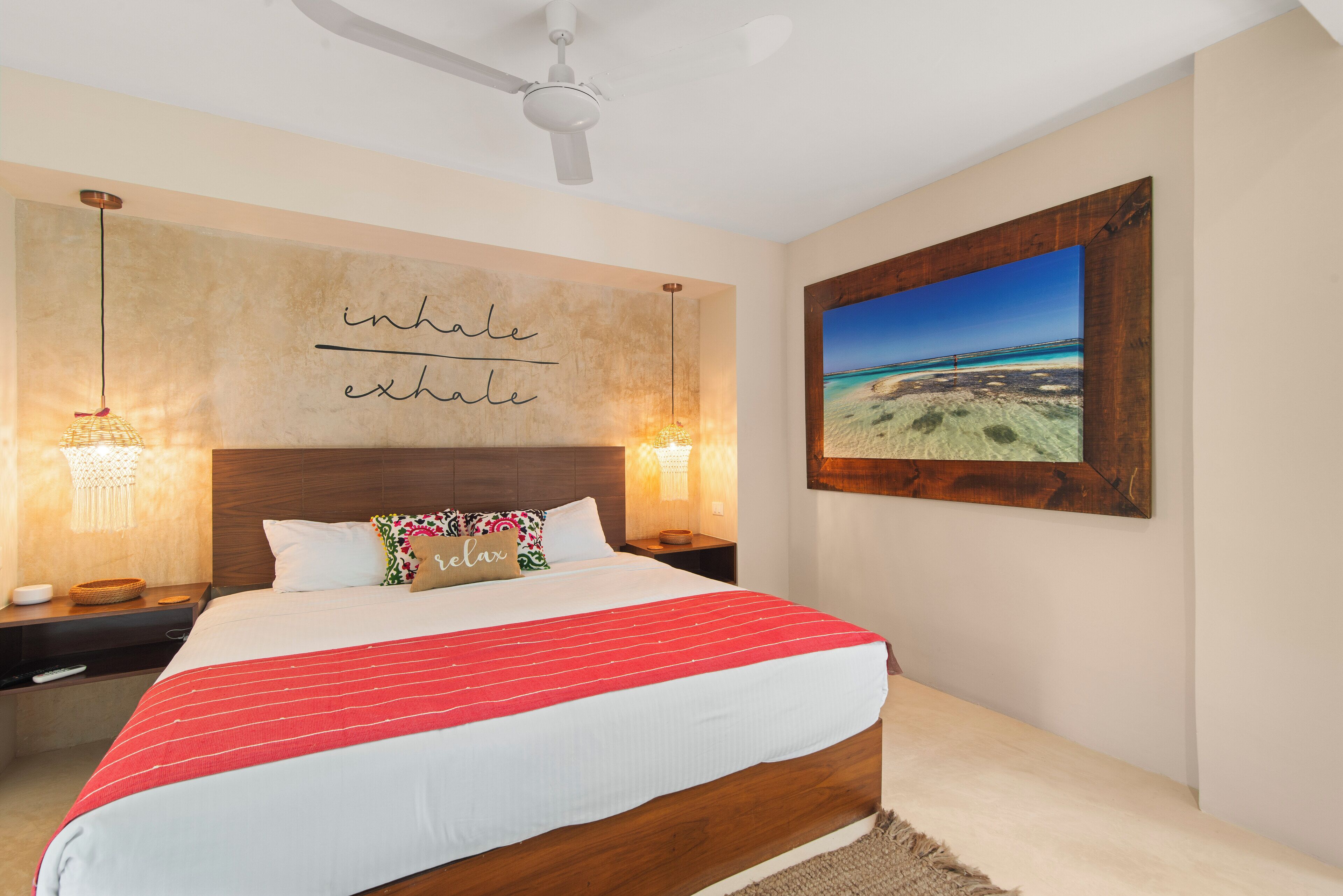 A bedroom with a large bed, a ceiling fan, a wall-mounted artwork, and a bedside table with a lamp.