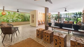 Amplia sala de estar con techo alto, ventiladores de techo y vista a exuberante vegetación. Hay taburetes de madera, un sofá y una mesa de comedor con sillas.