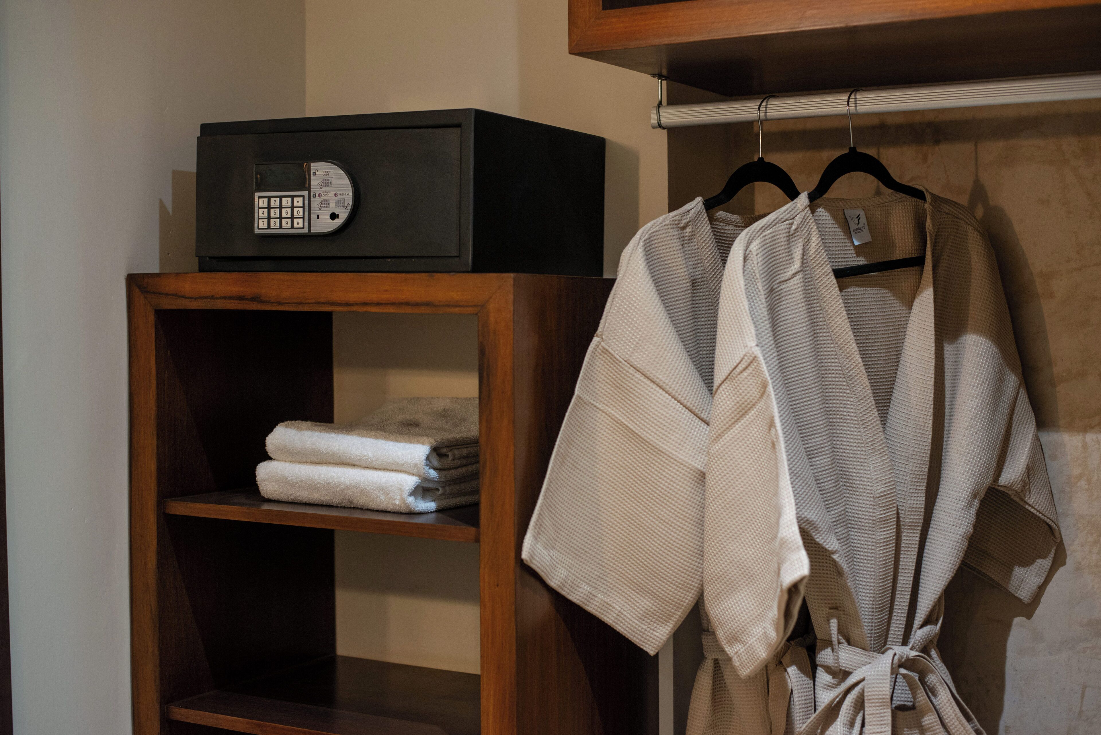 Une étagère en bois avec des serviettes pliées, un coffre-fort noir et un peignoir suspendu à un porte-manteau.
