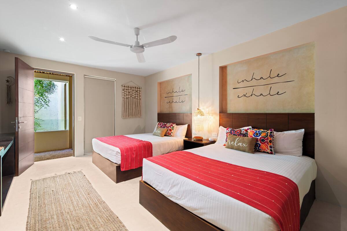 A hotel room with two beds, a ceiling fan, a large window with a view, and a motivational quote on the wall.