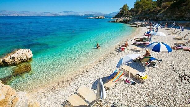 A pebble beach with clear turquoise water, sun loungers, and umbrellas.