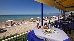 A beachside restaurant with outdoor seating overlooking the sea, featuring umbrellas and tables with food and drinks.