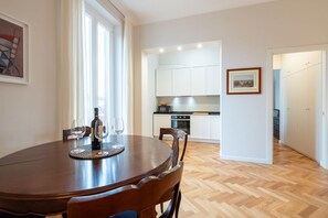 Un coin repas avec une table ronde, des chaises et une bouteille de vin. On aperçoit au fond une cuisine avec des meubles blancs et une cuisinière.