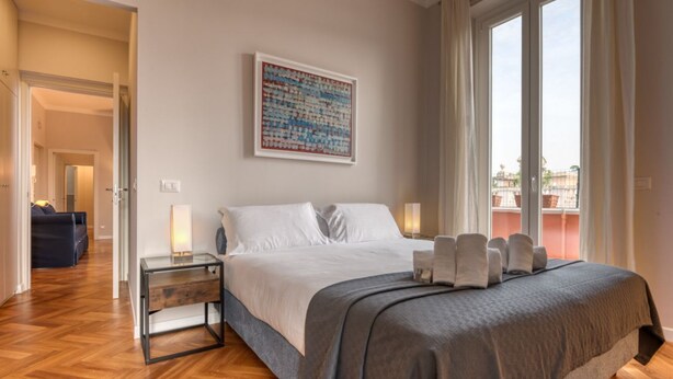 A hotel room with a bed, a nightstand, a lamp, a painting, and a view through the window. - Top view luxury apartment Piazza del Popolo (Roma)