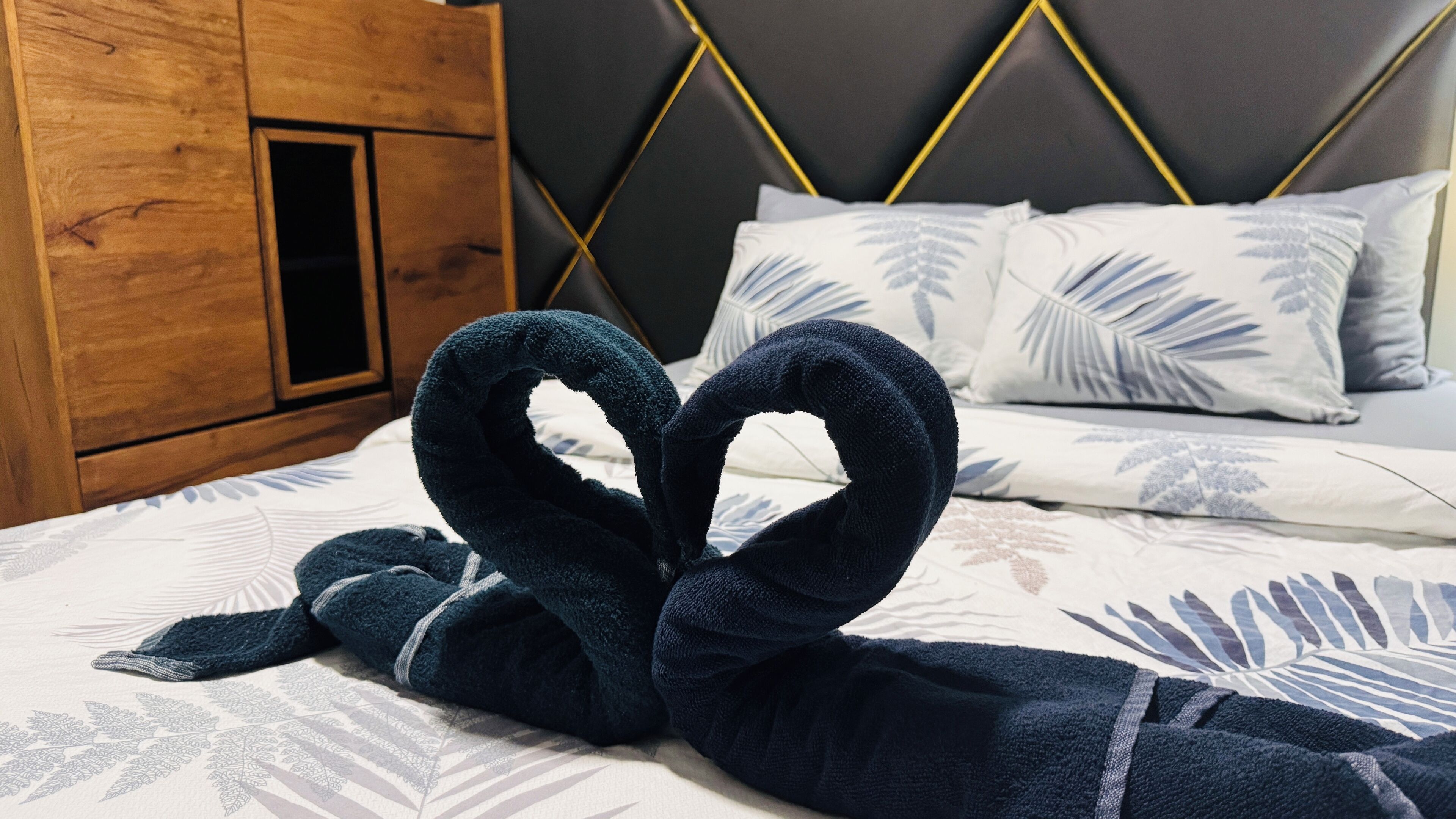 A bed with a dark blue towel folded in a heart shape, a wooden wardrobe, and a bed with white bedding and leaf-patterned pillows.