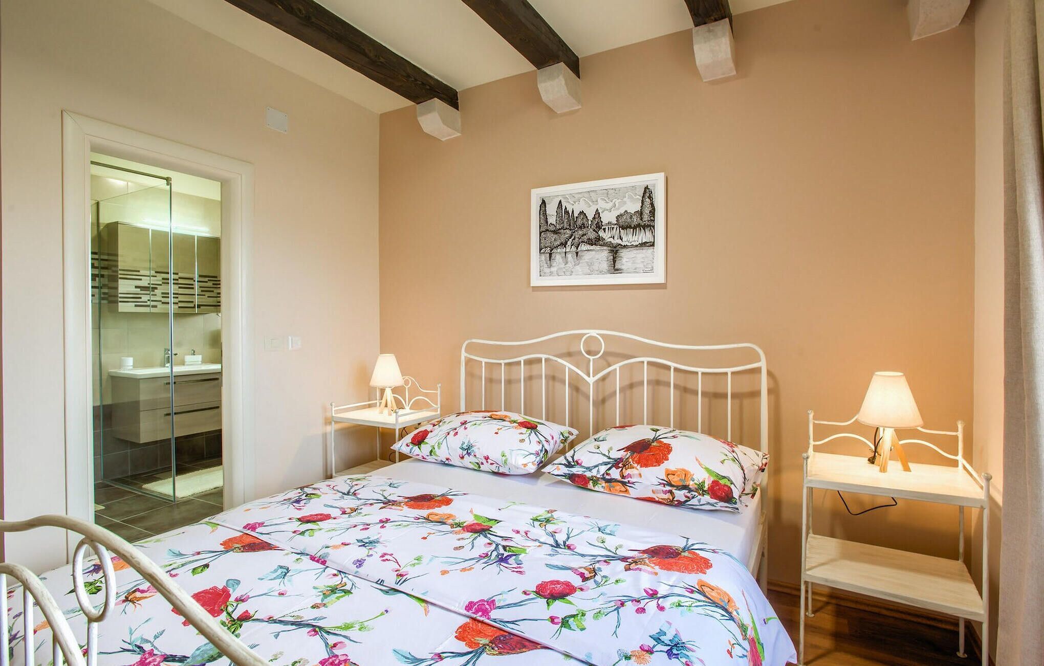 Een slaapkamer met een bed, twee nachtkastjes met lampen, een bijzettafeltje, een ingelijst beeld aan de muur, en een badkamer met een douchecabine van glas.