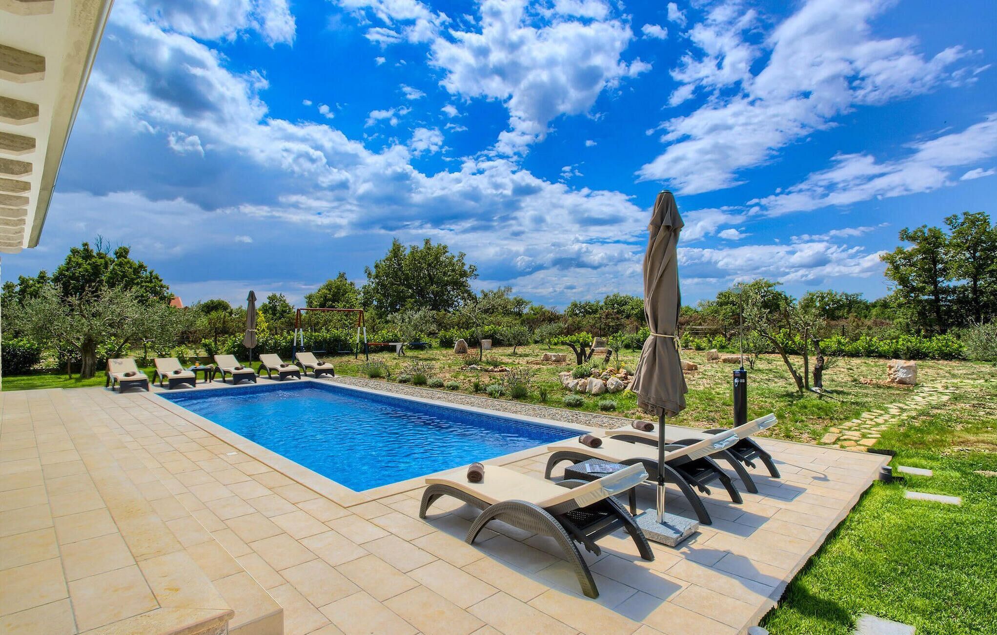 Ein Swimmingpool mit Liegestühlen und Sonnenschirmen, umgeben von einem Garten und Bäumen.