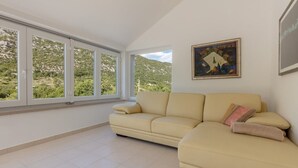Ein geräumiges Wohnzimmer mit einem beigefarbenen Sofa, großen Fenstern mit Blick ins Grüne und einem gerahmten Gemälde an der Wand.