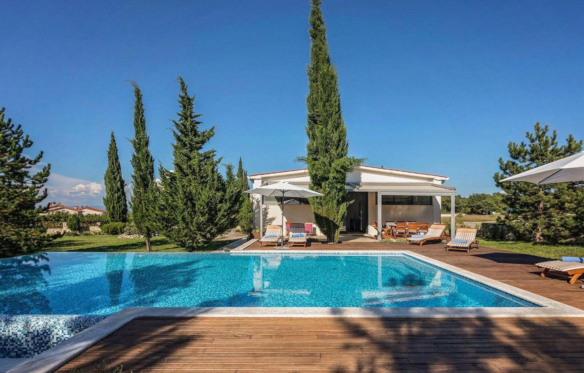 Una casa moderna con piscina, sillas de descanso y altos árboles.