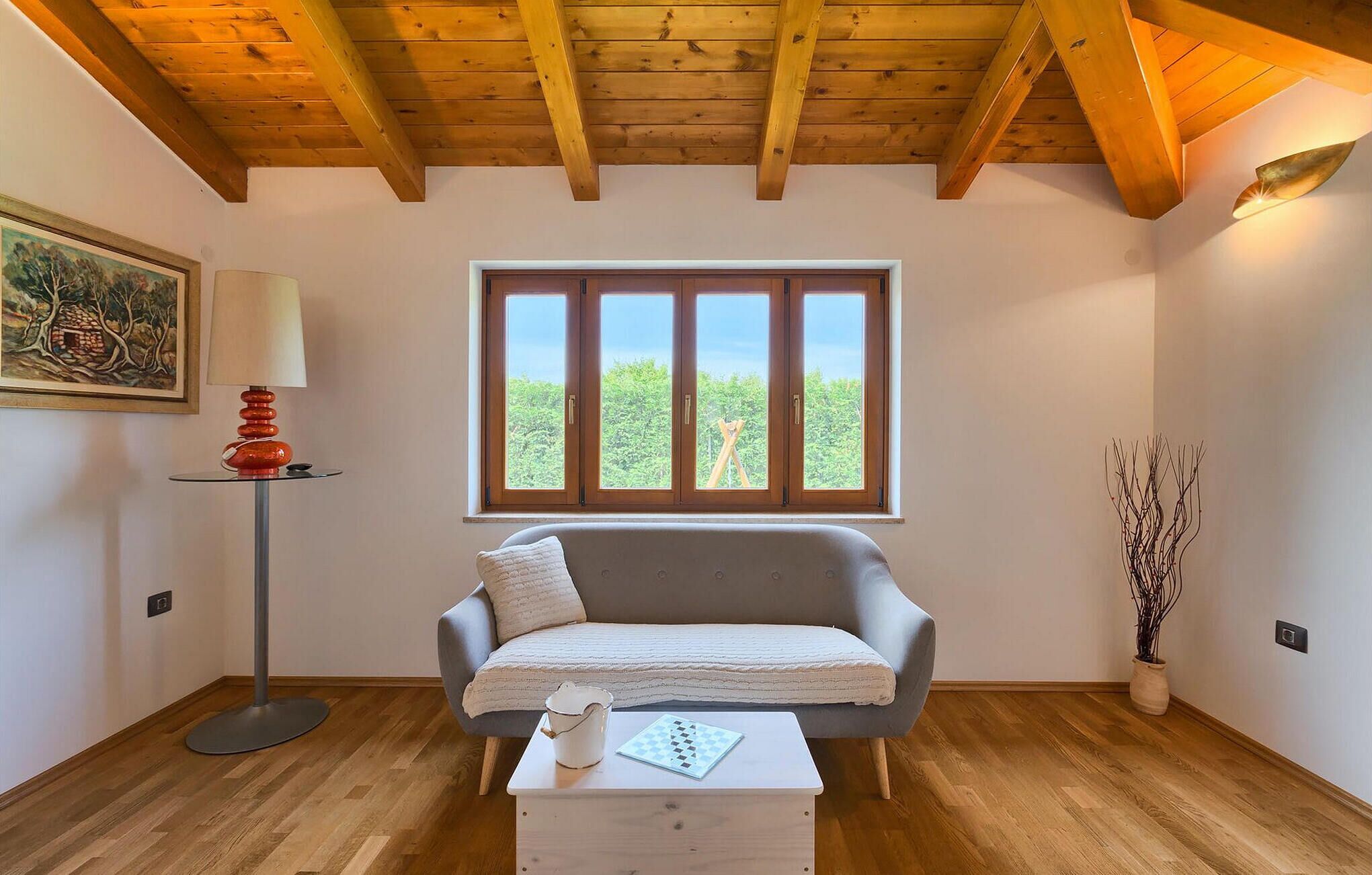 Ein gemütliches Zimmer mit Holzbalkendecke, einem grauen Sofa, einem kleinen weißen Tisch mit Schachbrett und einem Fenster mit Blick ins Grüne.