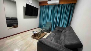 A living room with a sofa, a chair, a flat-screen TV, a clock, and a window with curtains. - Queen's Two Bedroom Apartment (Paramaribo)
