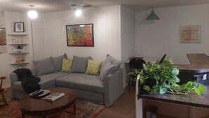 A living room with a grey sofa, a coffee table, a bookshelf, a framed picture, and a kitchen area with a plant. - 1 bedroom basement suite, private entrance, safe,quiet ,tree lined neighborhood. (Sherwood Park)
