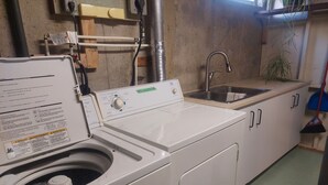 A laundry area with a front-loading washing machine, a sink, and a broom. - 1 bedroom basement suite, private entrance, safe,quiet ,tree lined neighborhood. (Sherwood Park)