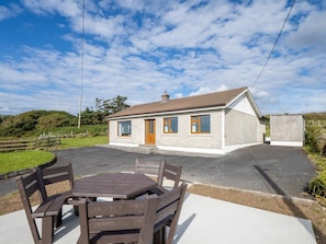 A single-story house with a paved driveway, a wooden table and chairs in the foreground, and a clear sky.