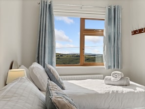 A bedroom with a bed, pillows, a lamp, a window with curtains, and a view of a landscape. - Mourneen Cottage with Sea Views (Cleggan)
