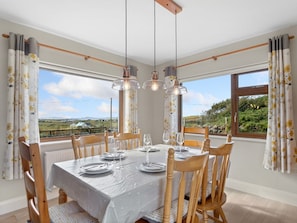 A dining area with a table set for four, wooden chairs, and a view of the countryside through large windows. - Mourneen Cottage with Sea Views (Cleggan)