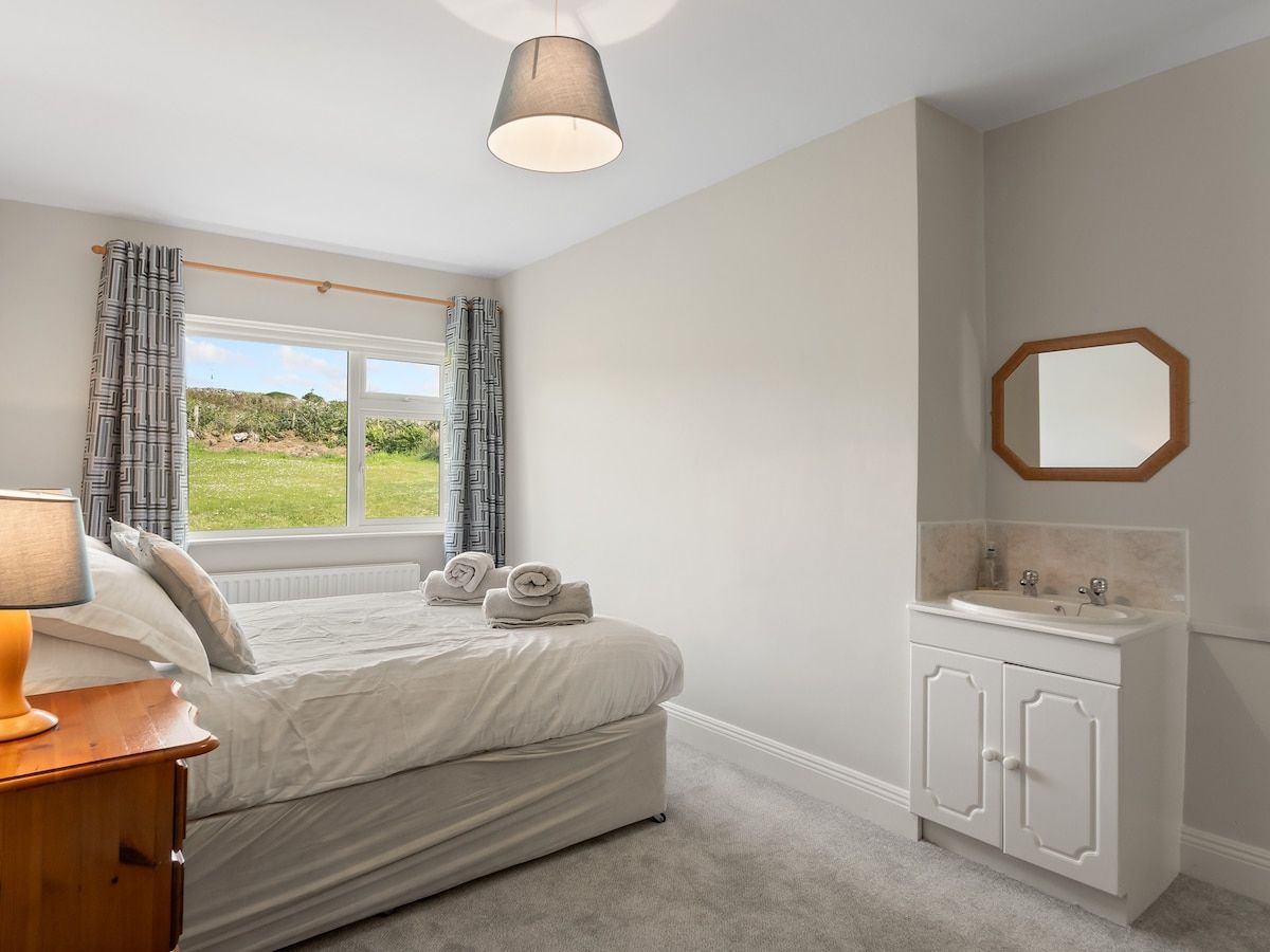 A bedroom with a bed, a bedside table with a lamp, a window with curtains, a bathroom with a sink and mirror.