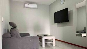 A modern living room with a grey sofa, a small white table, a mounted TV, and a wall-mounted air conditioner. - Queen's Two Bedroom Apartment with Pool View (Paramaribo)