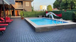 A swimming pool with a tiled deck, red lounge chairs, and a hedge with a red and white design. - Queen's Two Bedroom Apartment with Pool View (Paramaribo)