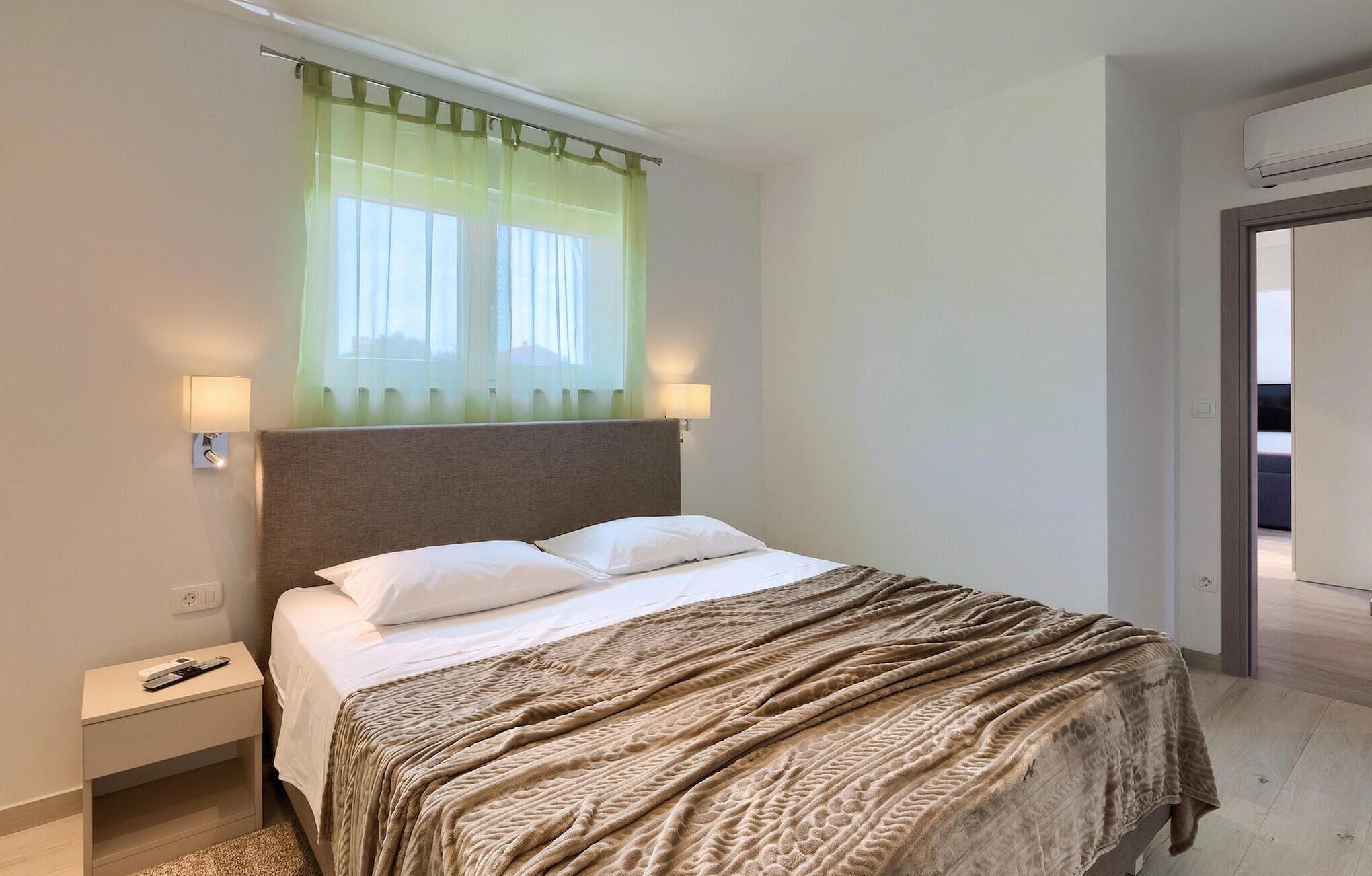 Een hotelkamer met een bed, een nachtkastje, een raam met dunne gordijnen en een deur die naar een andere kamer leidt.