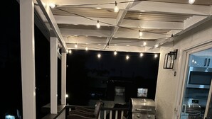 Un espacio al aire libre con patio, una zona de estar con cojines, una parrilla y una puerta de cristal que da a una cocina.