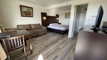 A hotel room with a wooden floor, a bed, a sofa, a desk with a chair, and a television.