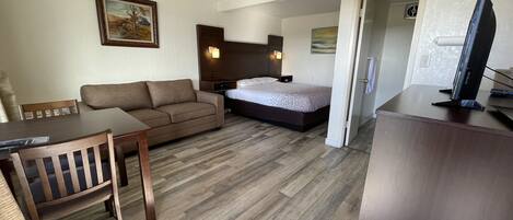 A hotel room with a wooden floor, a bed, a sofa, a desk with a chair, and a television.