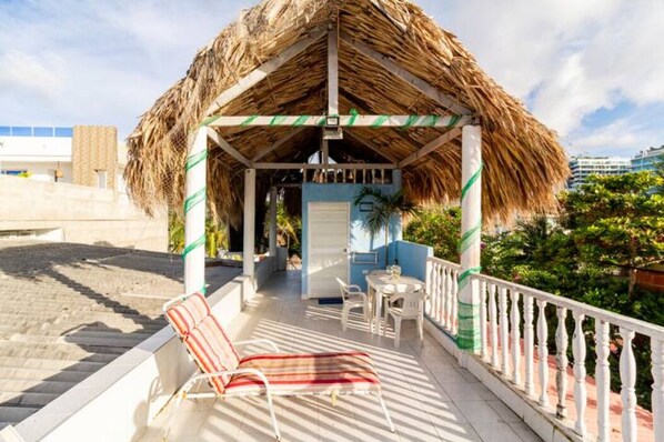 A covered outdoor seating area with a thatched roof, white railings, and a view of the beach and cityscape.