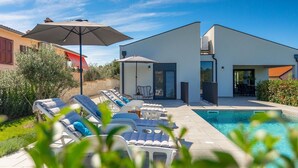 A modern outdoor pool area with lounge chairs and umbrellas, surrounded by a well-maintained garden and a two-story building. - Gorgeous home in Burici (Burici)