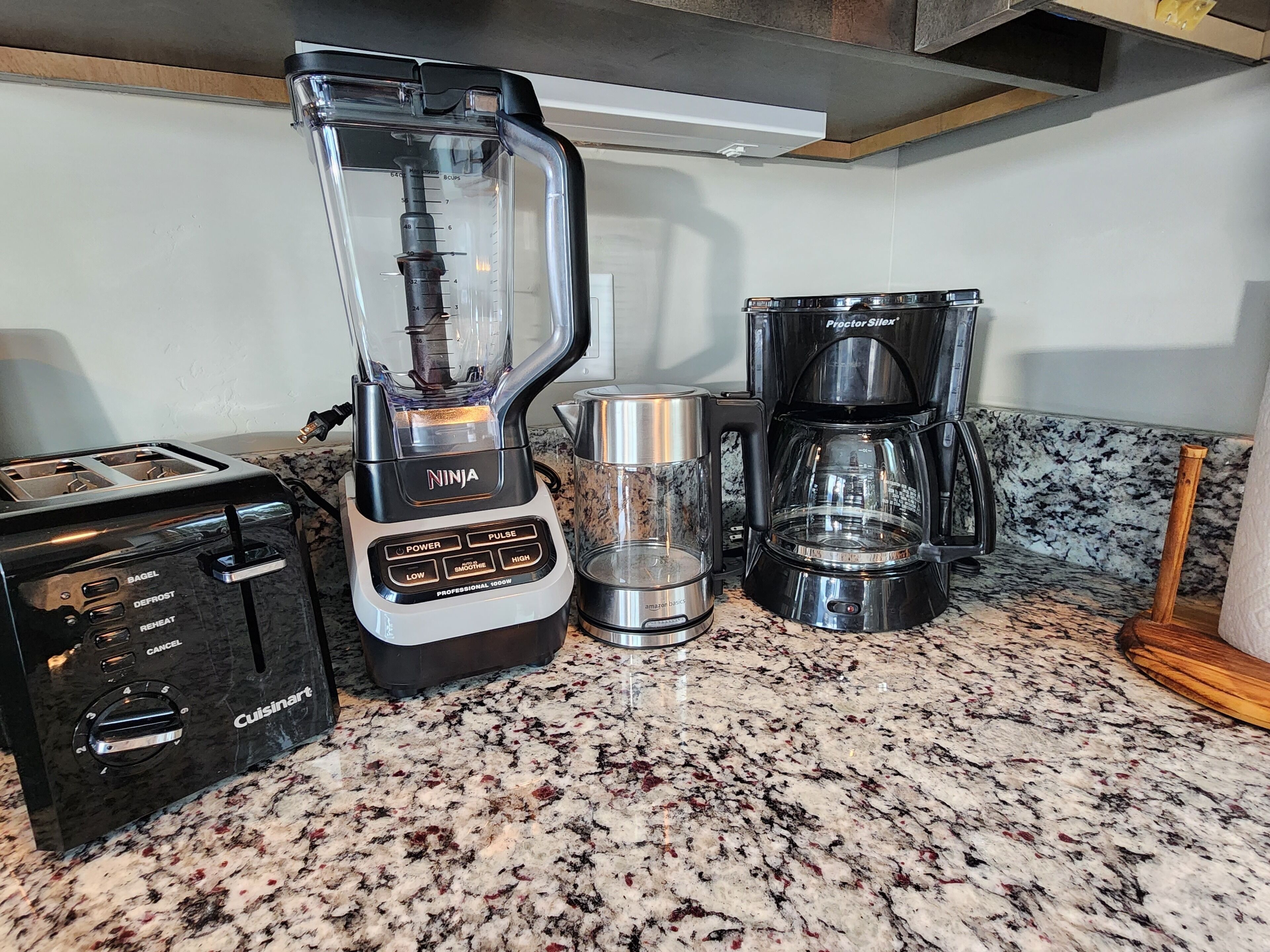 A kitchen with a Cuisinart toaster, a Ninja blender, a Proctor Sloane coffee maker, and a glass of water on a granite countertop.