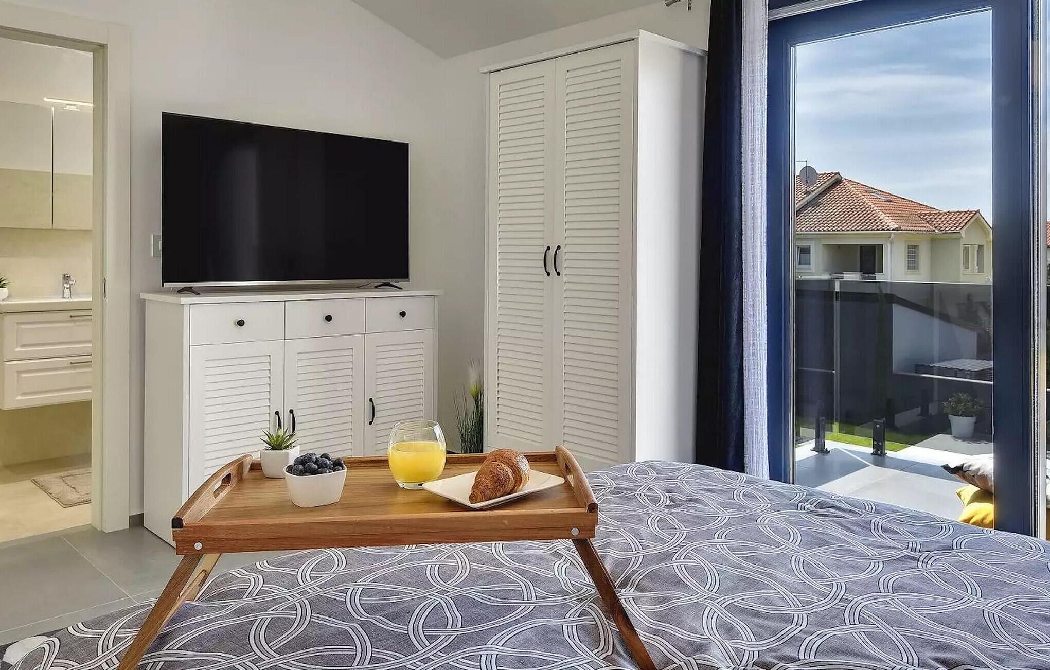 A bedroom with a bed, a TV, a wardrobe, a tray with breakfast, and a balcony with a view of houses.