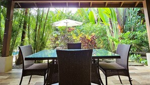 Une terrasse extérieure couverte, dotée d’une table et de chaises, entourée d’une végétation luxuriante et agrémentée d’un parasol blanc.