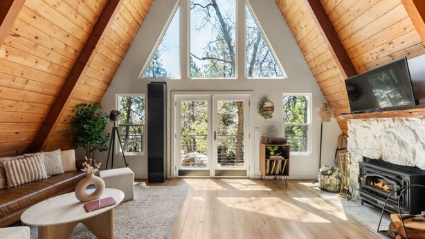 A cozy loft bedroom with a sloped ceiling, a fireplace, and large windows offering a view of the outdoors. - Luxury summer Scandia A-Frame mountain Chalet +Hot Tub, EV & Pet friendly (Running Springs)