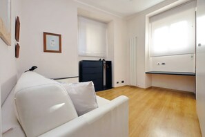 Une pièce avec un canapé blanc, une chaise noire, un bureau et une fenêtre avec des stores.