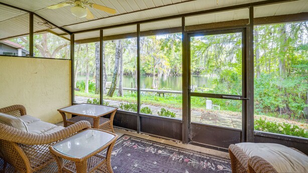 A covered outdoor seating area with wicker furniture and a glass table, overlooking a natural landscape with trees and a body of water. - Waterfront Dunnellon Retreat w/ River Access! (Dunnellon)