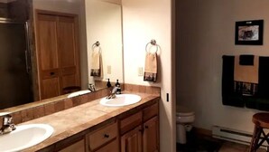 Un baño con dos lavamanos, un espejo y un inodoro. Hay toallas colgadas en ganchos y un cuadro en la pared.