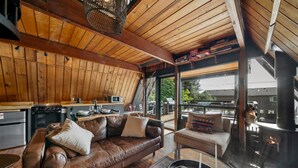 A cozy cabin interior with a wooden ceiling, a leather sofa, a dining area, and a wood-burning stove.