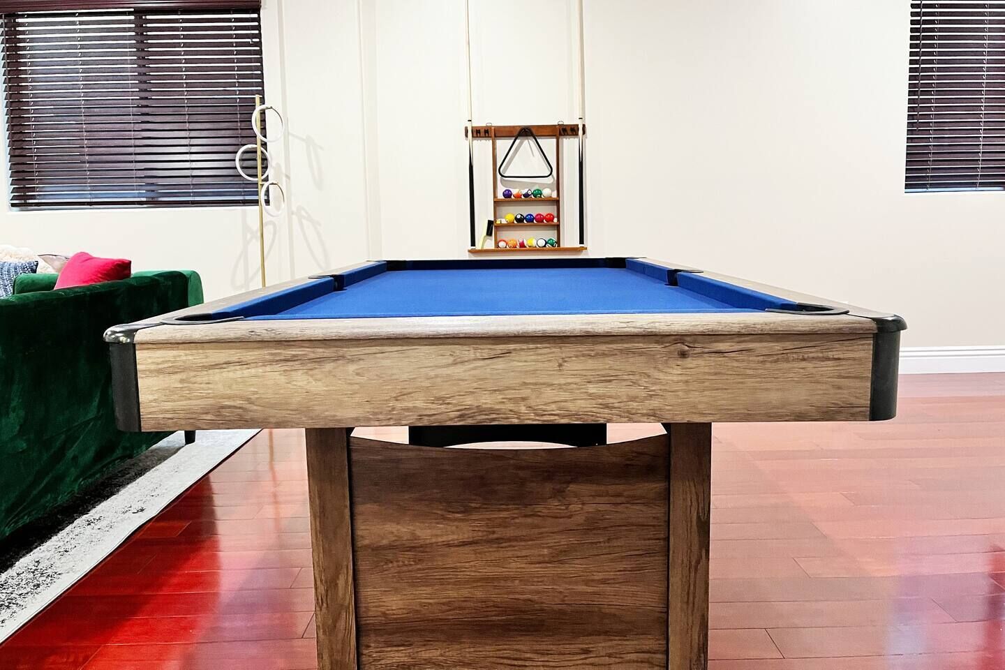 Een biljarttafel met houten frame en een blauwe viltkleed, geplaatst in een kamer met houten vloeren en een groene bank.