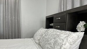 Un llit ben fet amb un llençol de color blanc i un coixí estampat, una tauleta de nit de fusta fosca amb un calaix, i un vas amb una flor blanca.