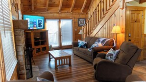 A cozy log cabin interior with a wooden staircase, a television mounted on the wall, a sofa with decorative pillows, and a wooden coffee table.