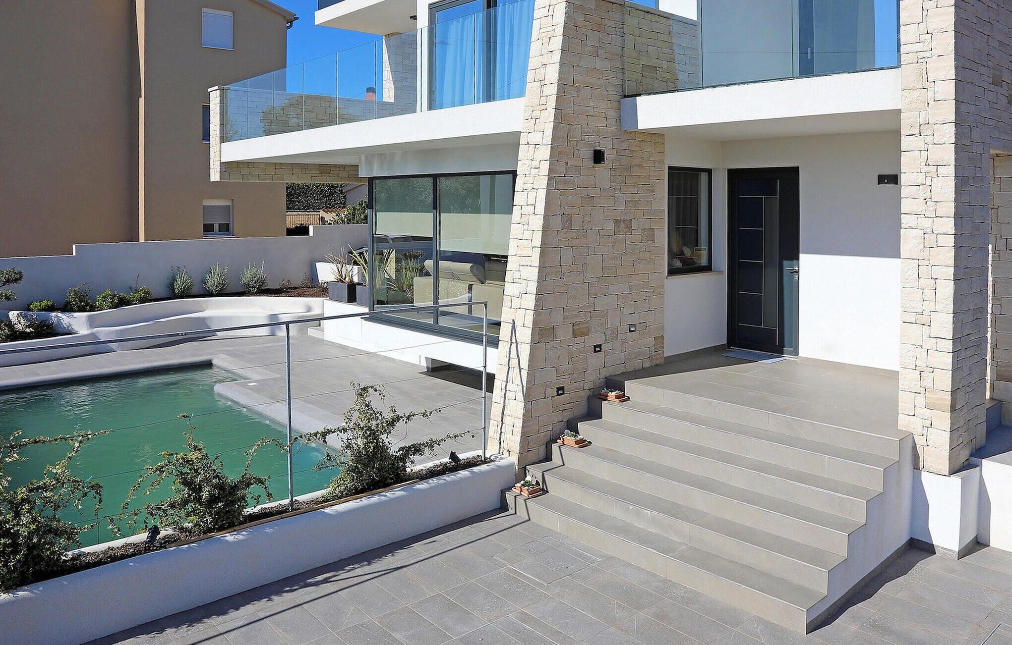 Une maison moderne à deux étages, dotée d’une piscine, d’une façade en pierre et d’un balcon en verre.