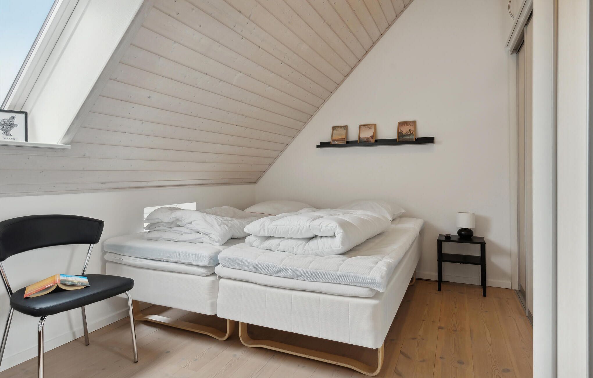 Un dormitorio con techo inclinado, cama con ropa blanca, una silla con un libro, mesita de noche con lámpara y estante con tres objetos decorativos.